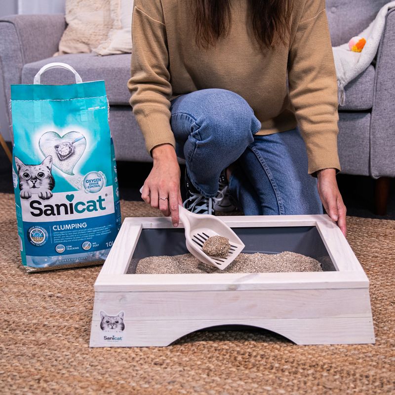 Sanicat Clumping cat litter with Oxygen Control, woman demonstrating use with scoop in wooden litter box. Packaging visible: 10L, antibacterial, dust-free.