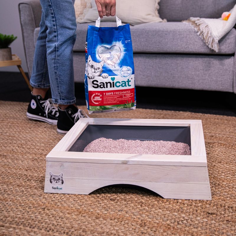 Person holding a bag of Sanicat litter above a filled litter tray, demonstrating use of the product for maintaining cleanliness.