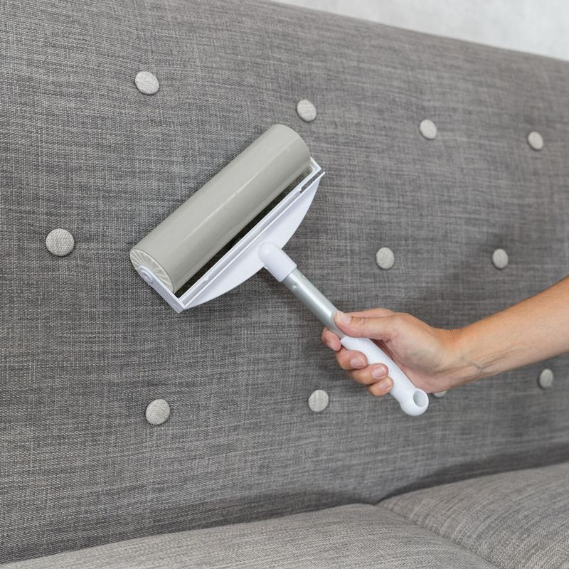 Hand holding a grey lint roller with handle, removing pet hair from a grey upholstered sofa.