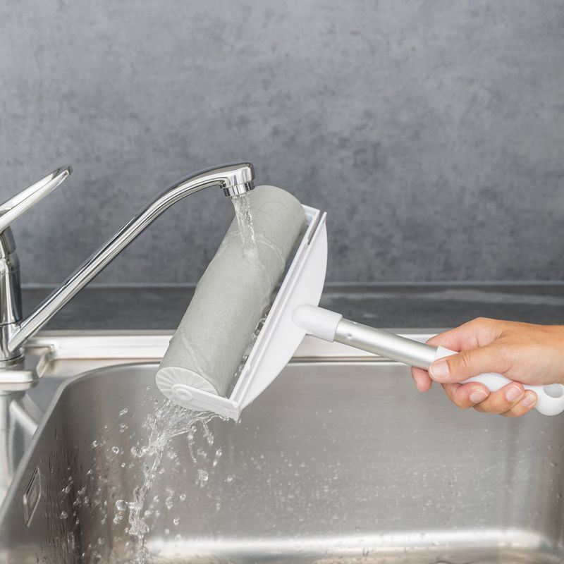 Hand holding a lint roller with washable design under running water at the sink. Shows easy cleaning of the roller.