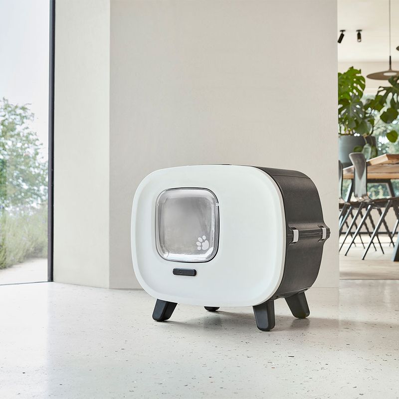 Modern cat house with white front, transparent door with paw motif, and black sides. Stands on four legs in a bright room.