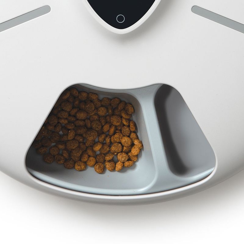 Automatic feeder with dry food in a grey bowl, second bowl empty. White housing with black control panel visible.