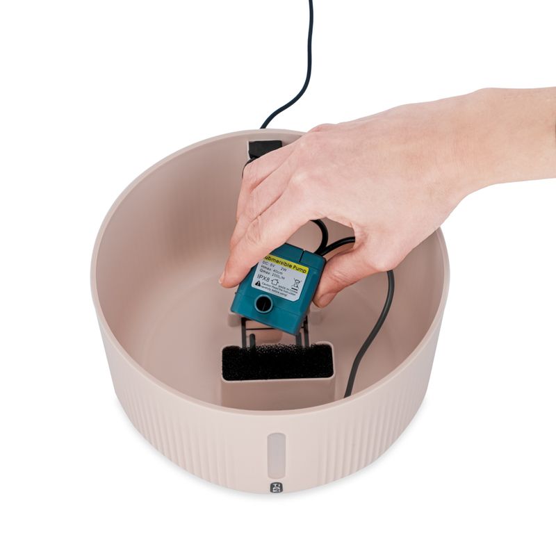 Hand showing installation of a submersible pump in a beige drinking fountain with filter insert.